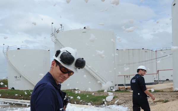 Two workers in navy blue uniforms and white hard hats are standing at an industrial tank farm facility, surrounded by large amounts of white firefighting foam dispersed in the air and on the ground. Large white cylindrical storage tanks are visible in the background, labeled 10-7, 30-2, and 10-9, with a 'No Smoking' sign visible. The scene appears to depict a foam suppression system discharge, either during a drill, test, or emergency response.
