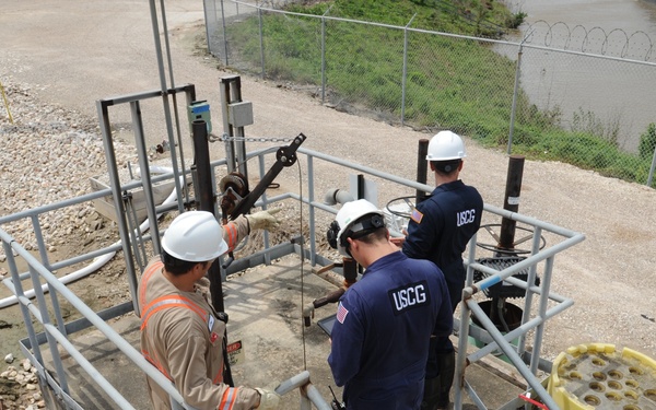Three workers are gathered on an industrial metal platform/grating structure inspecting or operating pipeline equipment near a waterway. Two individuals are wearing USCG (United States Coast Guard) uniforms in navy blue, while a third wears a tan coverall with a high-visibility orange vest. A fourth person in firefighting gear is visible in the lower right corner observing the activity.