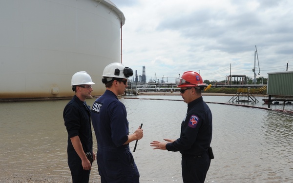 Three personnel in protective gear are conferring at what appears to be an industrial spill site, likely an oil or chemical facility. Two individuals wearing dark blue uniforms with 'USCG' markings and white hard hats appear to be U.S. Coast Guard members, while a third wears a red hard hat with a Texas-shaped patch. Containment booms are deployed in standing water near a large white storage tank, suggesting an active spill response operation.