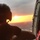 A person wearing a flight helmet is seated inside a military or rescue helicopter, looking out through the open door at a vivid sunset over a populated landscape below. The silhouetted figure and aircraft interior frame a dramatic orange and yellow horizon with the sun visible near the horizon line. The image captures an aerial perspective suggesting an active flight mission at dusk.