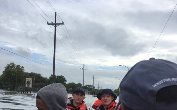 A flood rescue operation is underway, with multiple civilians being transported on a boat through heavily flooded streets. Two rescue personnel wearing orange life vests and dark caps are visible operating the vessel. A cap in the foreground reads 'HOUSTON RE...' suggesting this is likely related to Houston flood rescue operations, possibly during Hurricane Harvey (2017).