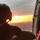 A person wearing a flight helmet is seated inside a military or rescue helicopter, looking out through the open door at a vivid sunset on the horizon. The silhouetted figure is backlit by warm orange and yellow tones from the low sun. Urban terrain is faintly visible far below, suggesting the aircraft is at significant altitude.