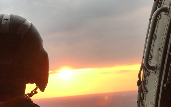 A person wearing a flight helmet is seated inside a military or rescue helicopter, looking out through the open door at a vivid sunset on the horizon. The silhouetted figure is backlit by warm orange and yellow tones from the low sun. Urban terrain is faintly visible far below, suggesting the aircraft is at significant altitude.