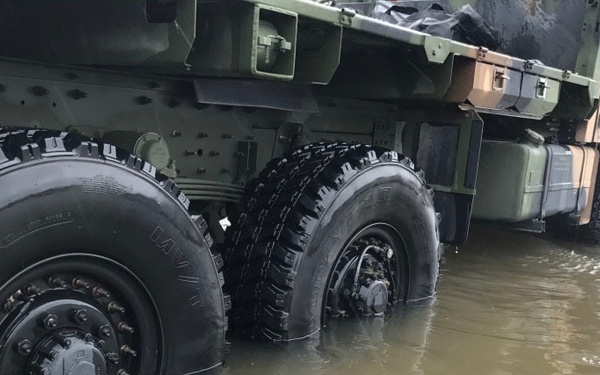 A military vehicle in camouflage paint is driving through heavily flooded terrain, with floodwater reaching approximately mid-wheel height on its large off-road tires. The vehicle appears to be a heavy tactical truck, likely engaged in flood relief or emergency response operations. The murky brown water and submerged road surface indicate significant flooding in the area.