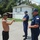 Three men are engaged in a conversation on a residential street. Two of the men are wearing navy blue uniforms consistent with the United States Coast Guard, including caps with insignia and American flag patches. The third man, shirtless with tattoos and wearing a baseball cap and sunglasses, appears to be speaking or gesturing to the uniformed personnel.