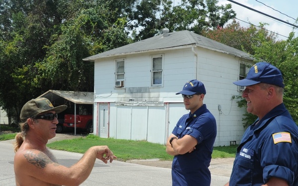 Three men are engaged in a conversation on a residential street. Two of the men are wearing navy blue uniforms consistent with the United States Coast Guard, including caps with insignia and American flag patches. The third man, shirtless with tattoos and wearing a baseball cap and sunglasses, appears to be speaking or gesturing to the uniformed personnel.