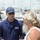 A U.S. Coast Guard member in uniform is engaged in conversation with a civilian at a marina. The USCG personnel member is wearing a navy blue uniform with 'USCG' lettering and a U.S. Coast Guard cap, speaking with an older man with long gray hair and a beard. Additional Coast Guard personnel and multiple docked boats are visible in the background.