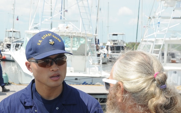 A U.S. Coast Guard member in uniform is engaged in conversation with a civilian at a marina. The USCG personnel member is wearing a navy blue uniform with 'USCG' lettering and a U.S. Coast Guard cap, speaking with an older man with long gray hair and a beard. Additional Coast Guard personnel and multiple docked boats are visible in the background.