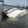 A vessel named 'Miss Kenzie' is capsized and partially submerged in a harbor or waterway, lying on its side with significant rust and deterioration visible on the hull. The boat appears to be secured with ropes or cables, possibly to prevent it from sinking further or drifting. Other working vessels and industrial infrastructure are visible in the background.