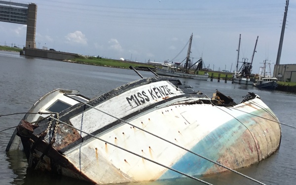 A vessel named 'Miss Kenzie' is capsized and partially submerged in a harbor or waterway, lying on its side with significant rust and deterioration visible on the hull. The boat appears to be secured with ropes or cables, possibly to prevent it from sinking further or drifting. Other working vessels and industrial infrastructure are visible in the background.