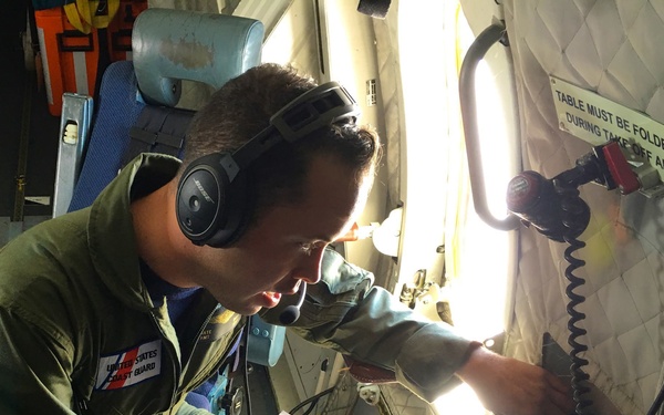 A United States Coast Guard crew member is seated inside a military aircraft, wearing a flight suit and Bose aviation headset with boom microphone. He is actively writing notes on a clipboard with paper, appearing to document information during what seems to be an active mission or patrol. The interior of the aircraft shows standard military aviation fixtures including a fold-up table, safety signage, and overhead lighting.