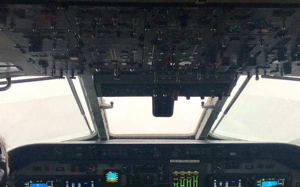 Two flight crew members are seated in the cockpit of a military aircraft, likely a WC-130 or similar reconnaissance/weather aircraft, flying through dense cloud cover or a storm system. The cockpit features a modern glass avionics suite with multiple multifunction displays, and the right-seat pilot is holding a tablet displaying weather radar imagery. The exterior visibility is near-zero, suggesting flight through clouds or a weather system.