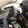 The image shows the interior of a military aircraft with multiple crew members in olive/flight suits working at an operational workstation. Personnel are wearing Bose headsets and US flag patches are visible on their uniforms, indicating US military affiliation. Crew members appear to be engaged in active mission operations, with one writing in a logbook and others monitoring equipment.