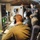 Three military personnel in olive/green flight suits are working inside what appears to be a military aircraft's mission systems compartment. They are wearing aviation headsets and operating multiple electronic workstations with green-lit displays and control panels. All personnel have visible U.S. flag patches on their right shoulders, indicating U.S. military affiliation.