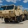 Hurricane Harvey Response A tan/desert-colored Oshkosh military heavy tactical truck (HEMTT or similar) is parked on a concrete surface, towing a lowboy trailer carrying a SkyTrak telescopic forklift. The scene appears to be a military staging or logistics area with other military vehicles visible in the background. The image was taken under clear blue skies with scattered clouds.