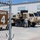 Hurricane Harvey Response A military installation showing a soldier interacting with large tan military trucks (likely HEMTT or similar heavy tactical vehicles) parked near a warehouse or hangar facility. A 'Gate 4' sign is visible on a chain-link fence in the foreground. A case of bottled water sits on the ground near the vehicles.