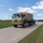 Hurricane Harvey Response A tan/sand-colored military tactical truck, consistent with an LMTV or FMTV (Family of Medium Tactical Vehicles), is traveling along a concrete road on what appears to be a military installation. The truck is towing a trailer and has a canvas-covered cargo bed. The setting features open green grass fields, light poles, and additional military vehicles visible in the background.