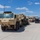 Hurricane Harvey Response A convoy of military heavy transport vehicles, primarily Oshkosh-manufactured trucks in desert tan livery, is staged on a large paved military installation. The lead vehicle is an Oshkosh HEMTT (Heavy Expanded Mobility Tactical Truck) with visible unit markings, hauling additional military equipment on a flatbed trailer. At least one uniformed service member is visible near the vehicles, suggesting staging or pre-movement inspection activity.