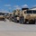 Hurricane Harvey Response A convoy of military vehicles in desert tan/camouflage paint is staged on a concrete apron, likely at a military installation or airfield. Several soldiers in combat uniforms are conducting pre-movement checks or coordinating the convoy lineup. A civilian white van is parked adjacent to the military vehicles on the right side.
