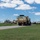 Hurricane Harvey Response A convoy of U.S. military tactical vehicles is moving along a paved road on what appears to be a military installation or base. The lead vehicle is a tan/desert-colored Heavy Expanded Mobility Tactical Truck (HEMTT) or similar 8-wheeled military logistics vehicle with headlights on. Additional military vehicles and equipment are visible in the background near a fenced compound area.