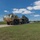 Hurricane Harvey Response A tan/desert-colored military heavy expanded mobility tactical truck (HEMTT) or similar 8-wheeled tactical vehicle is towing a lowboy flatbed trailer carrying a green military vehicle. The convoy is traveling on a paved road within what appears to be a military installation, surrounded by flat grassy terrain. The image was taken on a clear, sunny day with scattered white clouds.