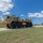 Hurricane Harvey Response A large tan/desert-camouflage military heavy tactical truck, consistent with an HEMTT (Heavy Expanded Mobility Tactical Truck), is traveling along a paved road on what appears to be a military installation. The vehicle is an 8-wheeled configuration towing or carrying additional equipment on its rear. The setting is a flat, open area with green grass, clear blue skies, and scattered clouds.