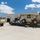 Hurricane Harvey A military logistics operation is underway at a Field Maintenance Shop facility. A tan/desert-colored telescopic handler (rough terrain forklift) is positioned on a flatbed trailer, likely being loaded or unloaded. A military HEMTT-style tactical truck is parked nearby, and personnel are observed directing or supervising the operation.