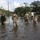 Several personnel in military/tactical attire and wading gear are walking through a flooded residential street. The floodwater reaches approximately knee to thigh depth, submerging the road and surrounding areas. The scene appears to be a post-disaster response operation in a suburban neighborhood.