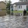 A flooded residential neighborhood shows standing water covering the street and yards to approximately knee depth. A person in military-style camouflage uniform and backpack wades through the floodwater toward a row of single-story homes. Parked vehicles and trash bins are partially submerged, indicating significant flood event impact.