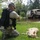 A military or National Guard soldier in camouflage uniform and tactical vest kneels beside a yellow Labrador Retriever lying on grass during what appears to be a flood rescue operation. A military LMTV (Light Medium Tactical Vehicle) is visible in the background partially submerged in floodwater, along with a partially submerged civilian car. The scene suggests a disaster response effort, likely following a hurricane or major flooding event.