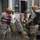 Military or emergency response personnel wearing yellow helmets, camouflage uniforms, and life vests are assisting civilians near a flooded residential property. A Texas-licensed vehicle is visible, and personnel appear to be handling a chain or tow equipment. The scene suggests a flood rescue or recovery operation, likely in Texas.