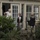 Two military or law enforcement personnel in camouflage uniforms and tactical vests are speaking with an elderly woman standing in the doorway of a house numbered 605. The scene appears to take place during or after a flooding event, as water is visible at ground level around the property. This appears to be a welfare check or evacuation assistance situation.