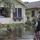 A flooded residential neighborhood shows standing water covering the yard and street in front of a yellow craftsman-style house. A person in military camouflage uniform carrying a backpack and holding an orange hose or rope stands in the floodwater, appearing to communicate with a civilian standing on the elevated front porch. An American flag is displayed on the house, and a white pickup truck is partially visible in the background.