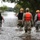 A group of five emergency responders are wading through a flooded street, with water reaching approximately knee to thigh height. The team includes military personnel, law enforcement, and rescue workers equipped with safety gear, suggesting an active flood rescue or search operation. The flooded road extends into the distance, with submerged vegetation and utility poles visible in the background.