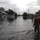 A flooded residential street shows emergency responders wading through floodwaters past single-story homes. At least three individuals wearing camouflage uniforms, orange life vests, and orange helmets are conducting what appears to be a search and rescue or damage assessment operation. The floodwater covers the entire street and appears to reach knee-to-thigh depth.