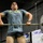 A muscular man is performing a barbell lift in a gym setting, appearing to be mid-movement during a deadlift or power clean. He is visibly sweating heavily, indicating intense physical exertion. The gym environment includes dumbbells, equipment, and motivational signage in the background.