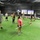A group of approximately 12-15 individuals are participating in a fitness training session inside an indoor turf facility. Most participants are holding white PVC pipes or lightweight bars, suggesting a weightlifting technique or movement drill class. The facility displays 'Storm Chasers' branding on banners in the background, and the space is equipped with various gym equipment along the walls.