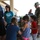 A group of children interact with a military working dog being handled by a soldier in camouflage uniform, likely at a community outreach or public relations event. Several adults, including at least two other uniformed military personnel, observe the interaction on what appears to be a covered outdoor patio or entrance area. The atmosphere appears friendly and informal.