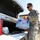 A U.S. Army soldier in ACU (Army Combat Uniform) is loading cases of bottled water into the trunk of a white vehicle with a Texas license plate. The scene appears to be a relief or supply distribution operation, likely in response to a disaster or emergency. Pallets of supplies are visible in the background.
