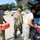 Three military personnel in camouflage uniforms are loading or unloading supplies into the open trunk of a dark-colored SUV in a parking lot. One soldier is carrying a packaged bag of what appears to be food or supplies with orange packaging, while another is handling water bottles near the vehicle. The setting appears to be a disaster relief or humanitarian aid distribution operation.