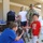 Two young boys interact with a gray/blue pit bull-type dog on a covered outdoor patio. A U.S. Army soldier in OCP camouflage uniform and boonie hat stands nearby, while adults in the background appear to be organizing supplies including boxed water and other goods. The setting suggests a community relief or aid distribution event.