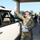 A U.S. Army soldier in OCP (Operational Camouflage Pattern) uniform is handing plastic bags through the window of a white pickup truck, appearing to distribute supplies or relief items. The scene takes place under a covered drive-through area with multiple civilians and volunteers also participating in what appears to be a relief distribution event. The setting and activity suggest a disaster relief or community assistance operation.