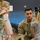 A U.S. Army soldier named Norkett, wearing OCP (Operational Camouflage Pattern) uniform with an American flag patch, is crouched down at child level interacting with several young children in what appears to be a classroom or childcare setting. The soldier appears to be engaged in a community outreach or school visit activity. The setting includes colorful wall murals and classroom furniture visible in the background.