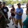 A U.S. Army soldier in camouflage uniform is engaged in conversation with a group of five civilians outside a building. The setting appears to be a military installation or community facility, with vehicles and palm trees visible in the background. The group appears to be in an informal discussion, possibly during a community outreach or relief effort.