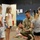 A U.S. Army soldier in camouflage uniform is crouching down and interacting with a group of young children in what appears to be a classroom or childcare facility. The soldier is engaged in conversation and play with the children, who are gathered around him. The setting features colorful murals on the walls suggesting a child-friendly educational environment.