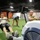 A group of individuals are engaged in a fitness training session inside an indoor sports facility with artificial turf flooring. Two men are prominently using Concept2 rowing machines in the foreground, while others perform various exercises in the background. The setting appears to be a multi-use athletic facility, possibly used for military or group fitness training.