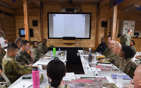 A military briefing or planning session is underway in a wood-paneled room, with approximately 20 uniformed personnel seated around a large conference table. A soldier standing at the front is presenting to the group, with a projected presentation visible on a screen behind him. Maps and documents are spread across the table, suggesting an operational or tactical planning meeting.