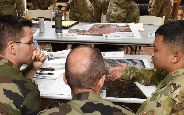 Three military personnel in different camouflage uniforms are seated around a table engaged in what appears to be a planning or briefing session. One soldier wearing US Army OCP (Operational Camouflage Pattern) uniform is pointing at maps or aerial imagery on the table, while two others in a different woodland camouflage pattern (consistent with French military uniform) listen and observe. The setting appears to be a joint multinational military planning environment with multiple documents, maps, and aerial photographs spread across the tables.