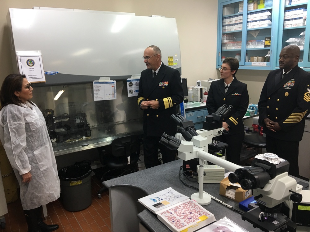Three U.S. Navy personnel in dress uniforms are visiting a laboratory, appearing to receive a briefing or tour from a civilian staff member wearing a white lab coat. The setting is a scientific laboratory identified as a Parasitology Department, equipped with microscopes, a biosafety cabinet, and supply storage. The interaction suggests an official inspection or command visit to a military medical or research facility.