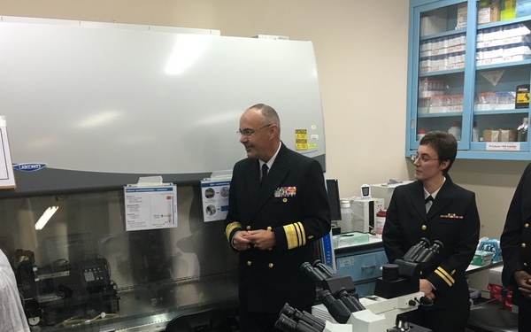 Three U.S. Navy personnel in dress uniforms are visiting a laboratory, appearing to receive a briefing or tour from a civilian staff member wearing a white lab coat. The setting is a scientific laboratory identified as a Parasitology Department, equipped with microscopes, a biosafety cabinet, and supply storage. The interaction suggests an official inspection or command visit to a military medical or research facility.